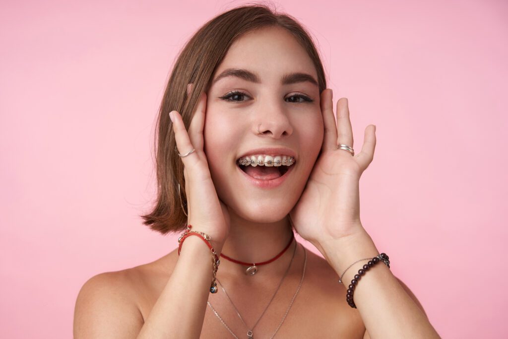 Cheerful young lovely brown-eyed brunette woman with short haircut holding her face with raised hands and smiling happily while posing over pink background