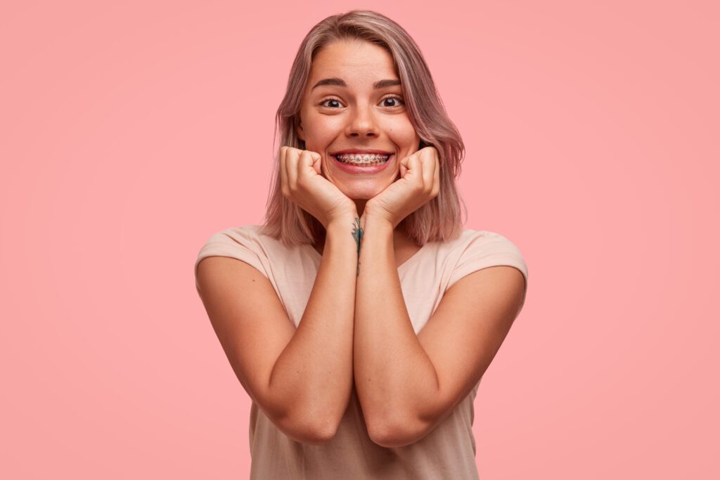 Glad satisfied woman with broad smile, keeps hands under chin, listens with joyful expression interesting story, expresses positive emotions, isolated on pink background. Facial expressions.